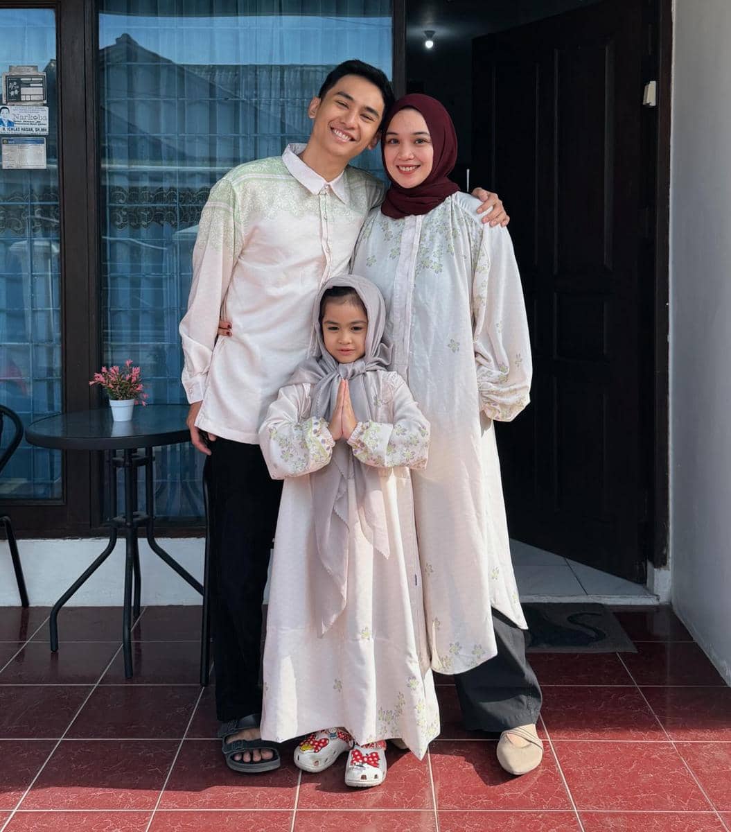 Seorang pria, wanita berhijab, dan anak perempuan kecil berpose bersama di depan rumah dengan pakaian senada berwarna pastel.