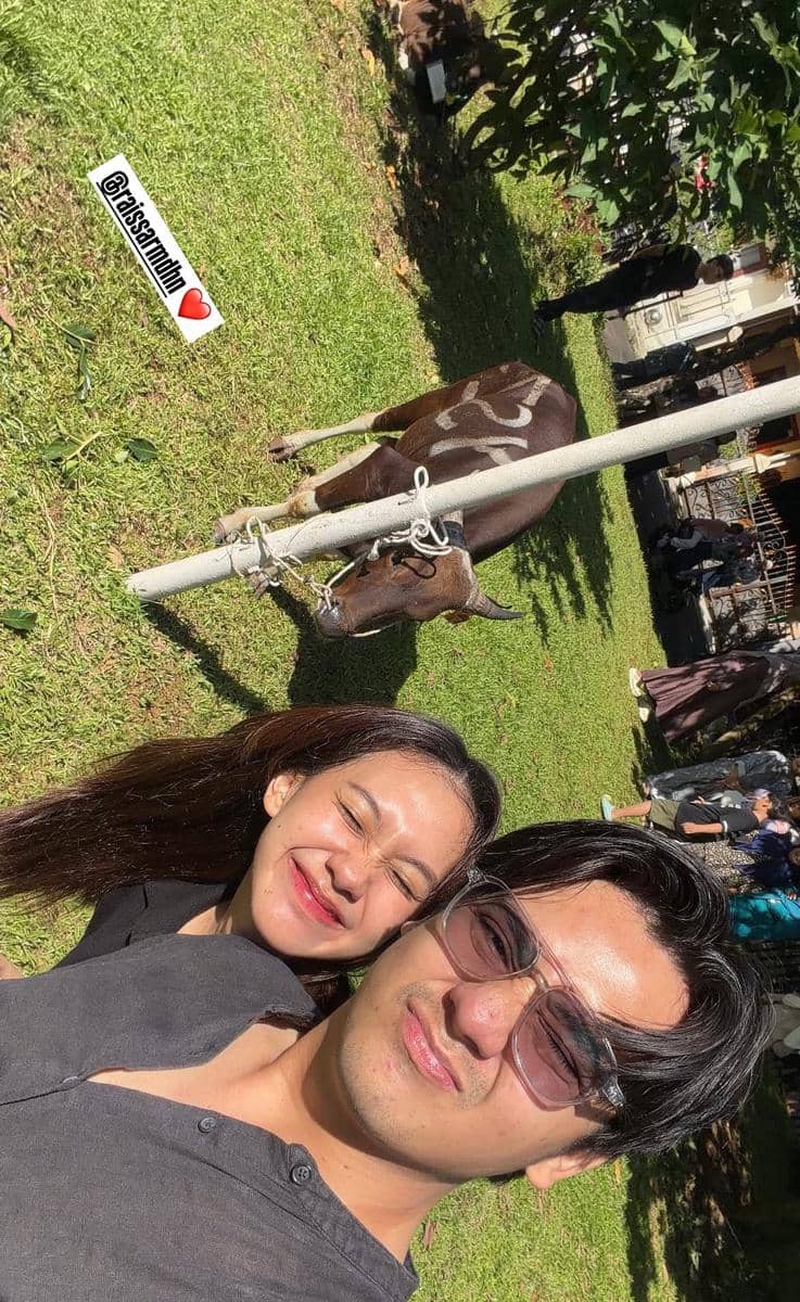 Sepasang orang berfoto selfie di bawah sinar matahari dengan seekor sapi kurban yang diikat di halaman rumput pada perayaan Idul Adha.