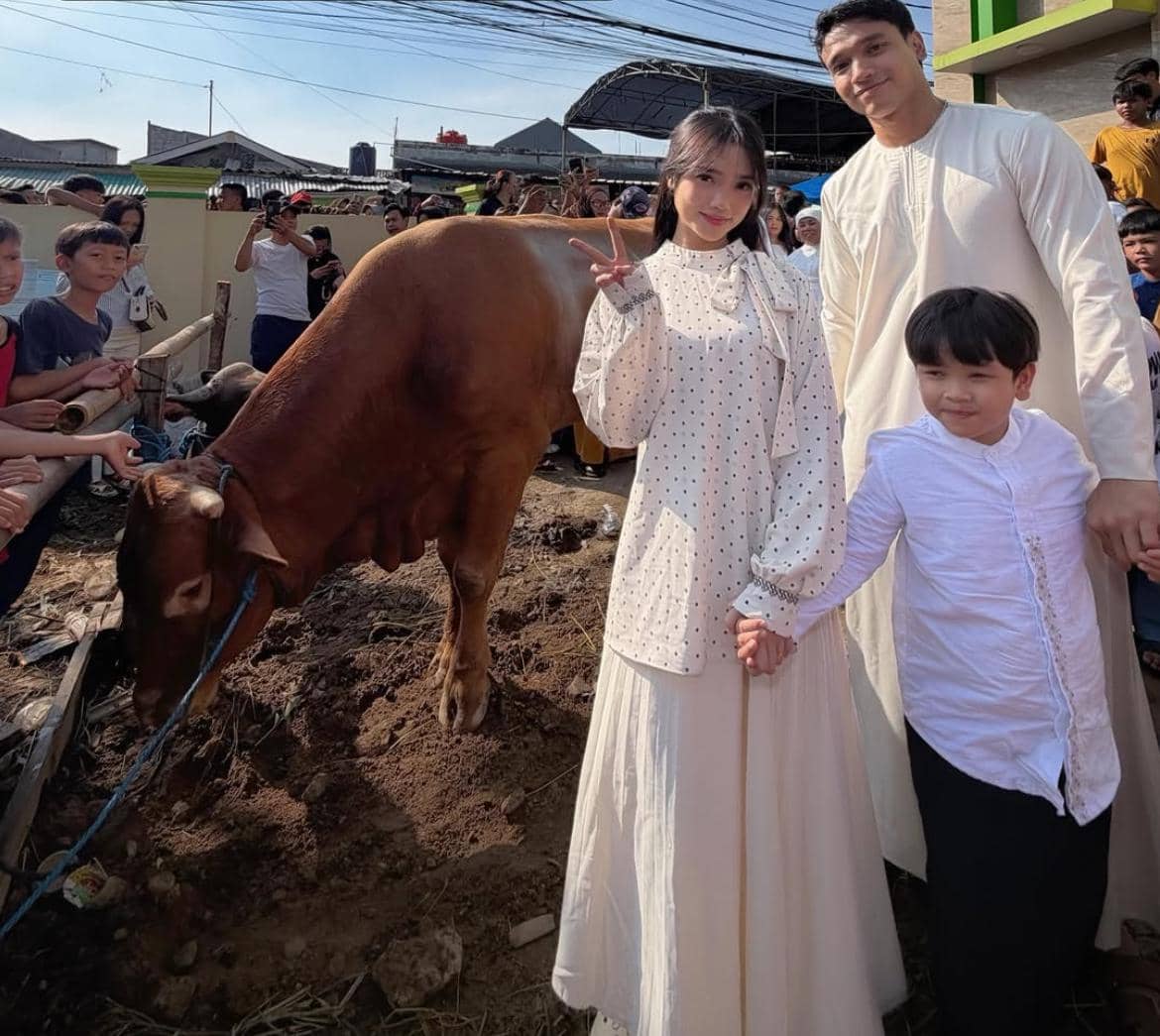 Seorang wanita, pria, dan anak kecil berbusana putih berpose di dekat sapi kurban saat perayaan Idul Adha 2026 di luar ruangan.
