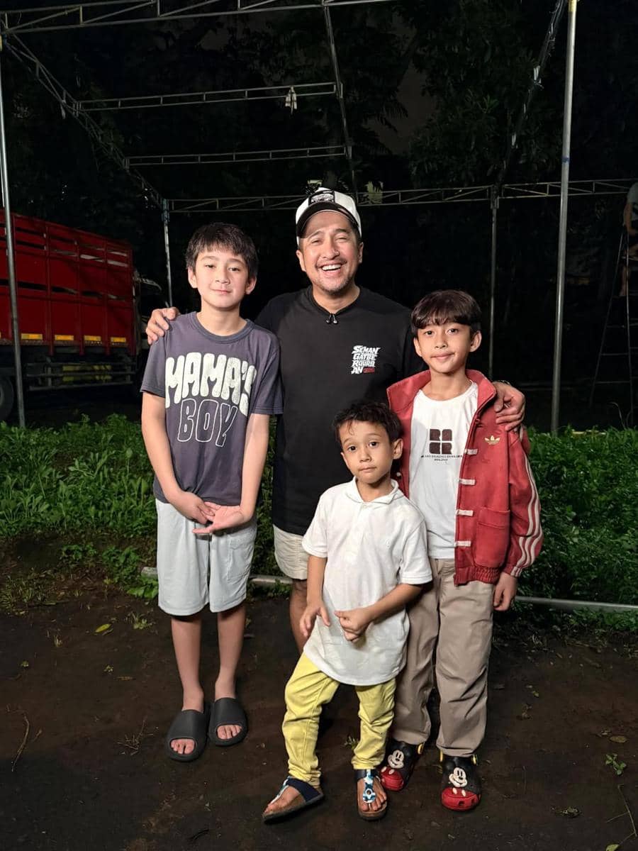 Seorang pria tersenyum berpose bersama tiga anak laki-laki di area luar ruangan pada malam hari dengan latar belakang hijau dan truk merah.
