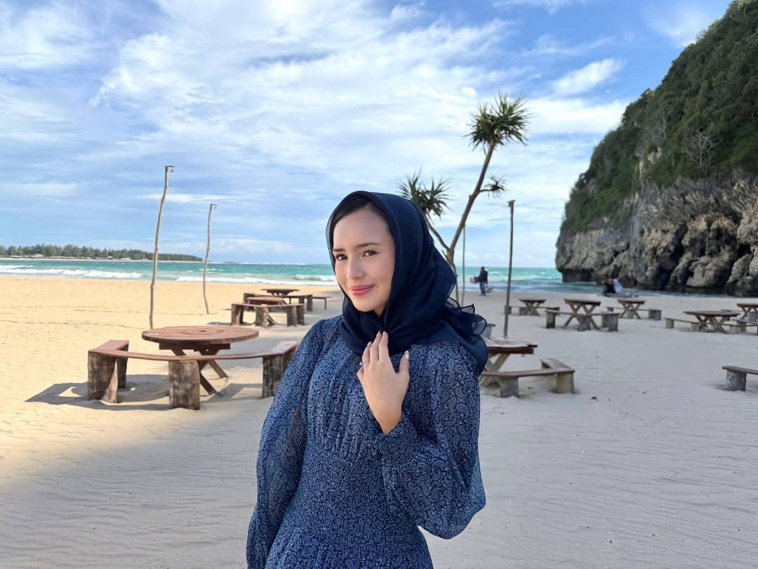 Seorang perempuan berhijab biru berdiri di pantai berpasir dengan latar laut biru, meja kayu, dan tebing hijau di kejauhan.