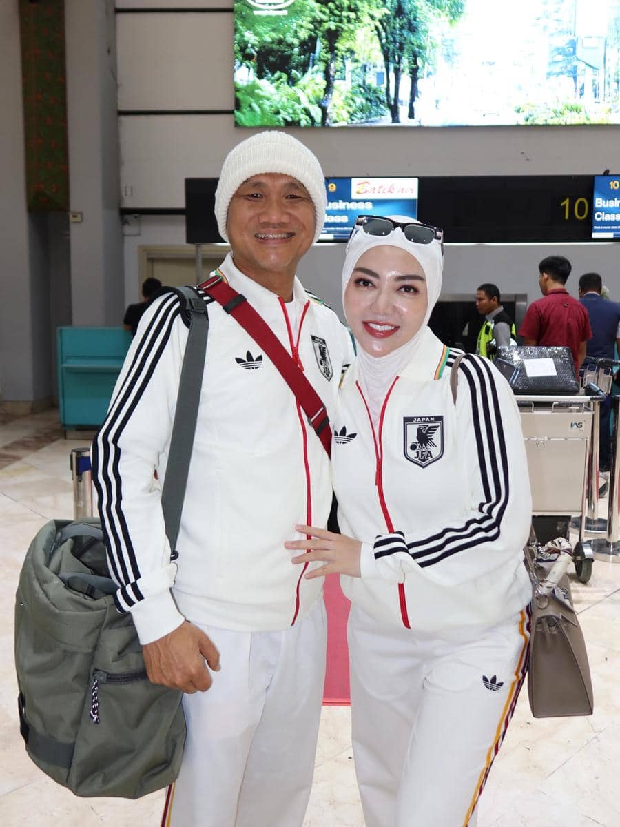 Sepasang pria dan wanita mengenakan jaket olahraga putih dengan garis hitam berpose di area check-in bandara sambil tersenyum ke kamera.