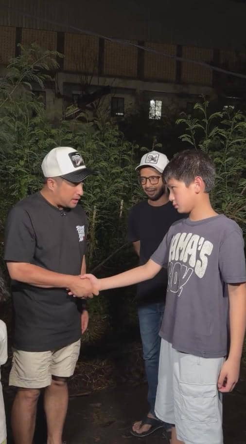 Seorang pria mengenakan topi putih dan kaus hitam berjabat tangan dengan anak laki-laki, disaksikan oleh pria lain di malam hari.