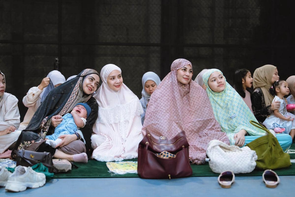 Sekelompok perempuan mengenakan mukena duduk bersama di atas sajadah saat perayaan Idul Adha, dengan suasana hangat dan penuh kebersamaan.