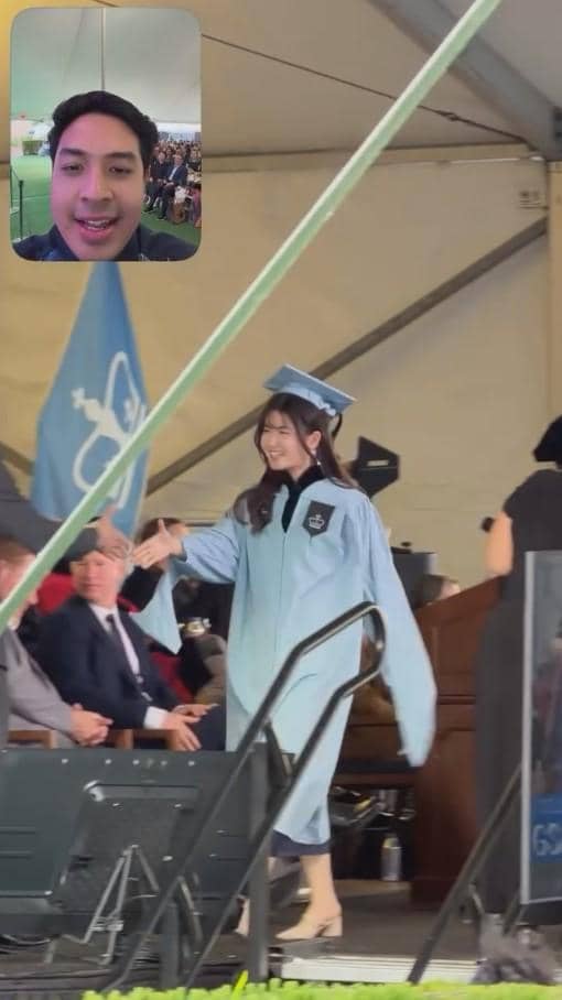 Seorang perempuan mengenakan toga wisuda berwarna biru muda berjalan di panggung sambil berjabat tangan, dengan panggilan video seorang pria di sudut gambar.