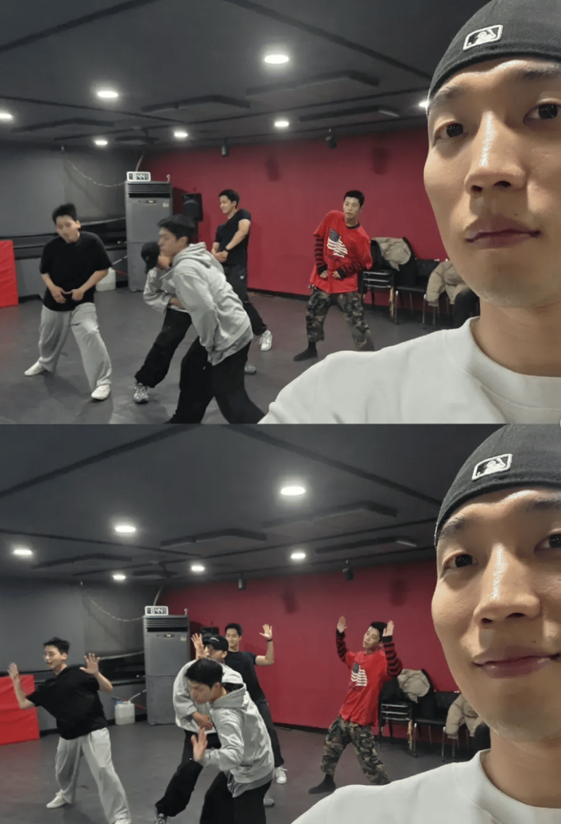 Beberapa anggota Migak Boys berlatih menari di studio dengan dinding merah, sementara satu orang mengambil swafoto di depan kamera.
