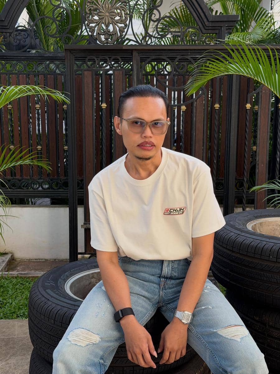 Seorang pria mengenakan kaus putih dan celana jeans robek duduk di atas tumpukan ban di area luar dengan pagar besi dan tanaman hijau di belakangnya.
