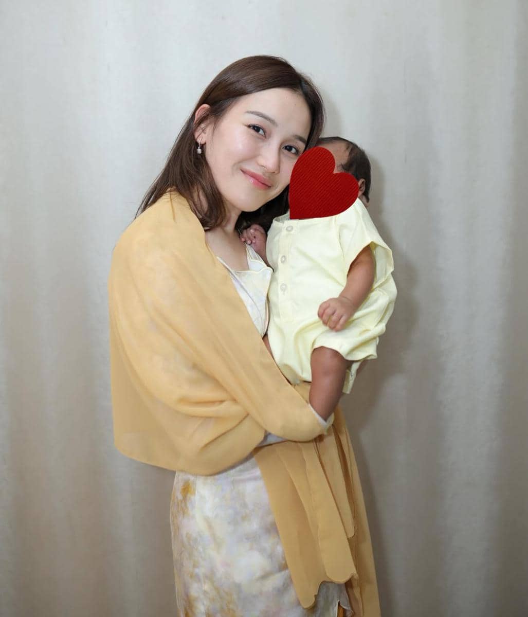 Seorang perempuan mengenakan selendang kuning lembut tersenyum sambil menggendong bayi berpakaian kuning di depan latar polos.
