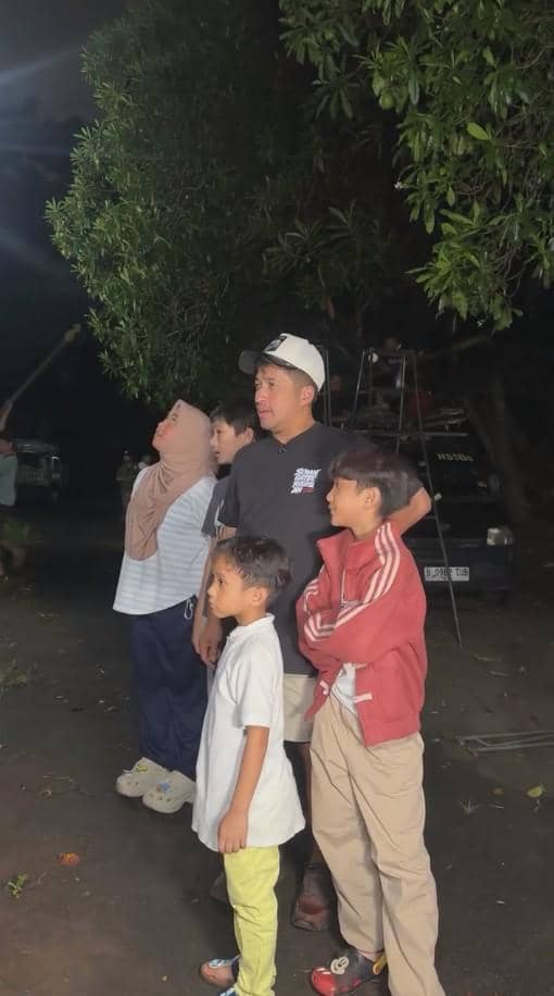Seorang pria bersama dua anak laki-laki dan seorang perempuan berhijab berdiri di luar ruangan pada malam hari sambil melihat ke arah atas.