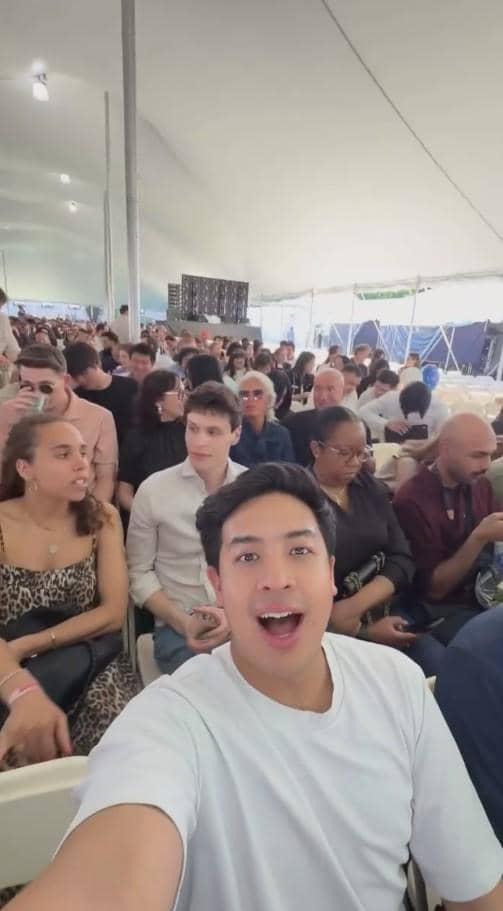 Seorang pria tersenyum sambil berfoto selfie di tengah kerumunan orang yang duduk di bawah tenda besar pada acara wisuda.