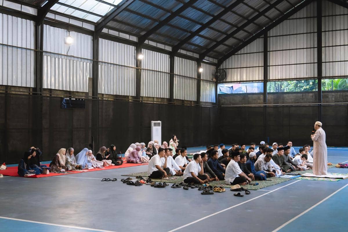 Seorang ustaz memimpin doa Idul Adha di dalam lapangan tertutup dengan jamaah pria dan wanita duduk terpisah di atas sajadah.