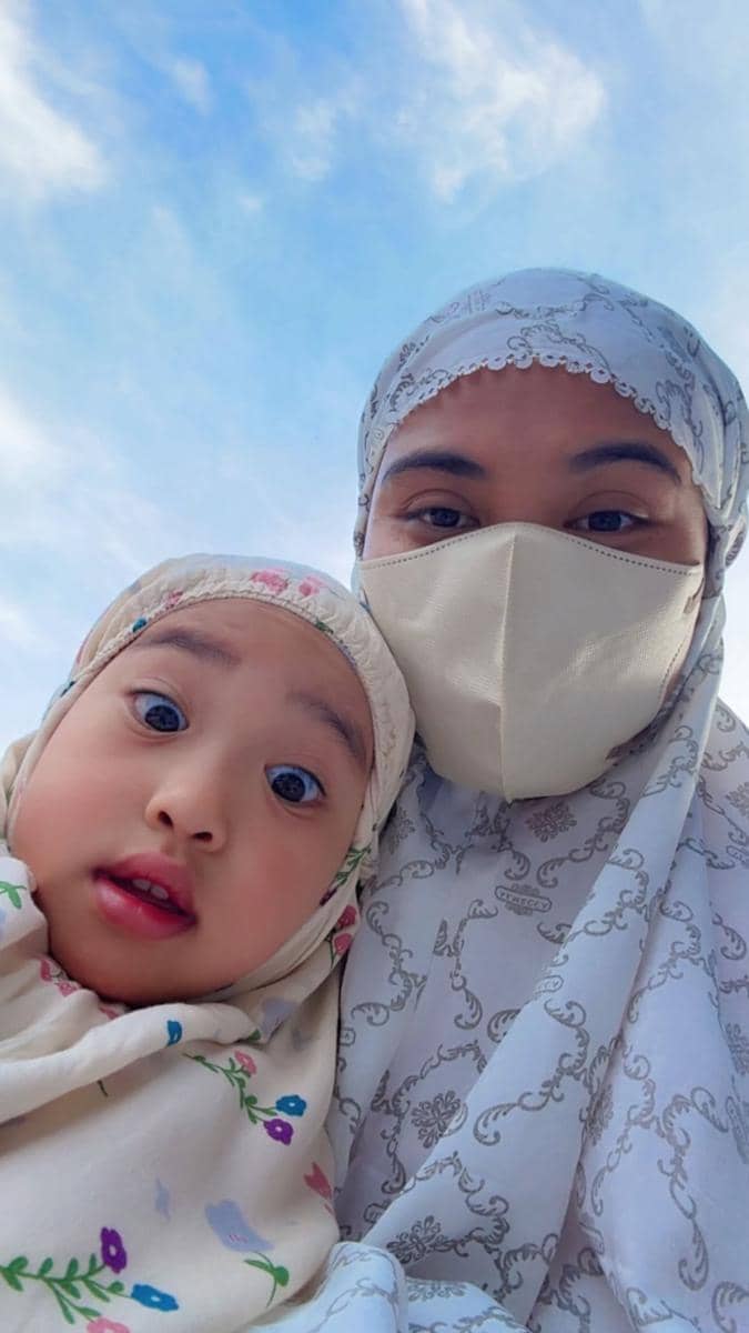 Seorang wanita berhijab dan bermasker berpose bersama anak kecil berhijab bermotif bunga dengan latar langit biru cerah.
