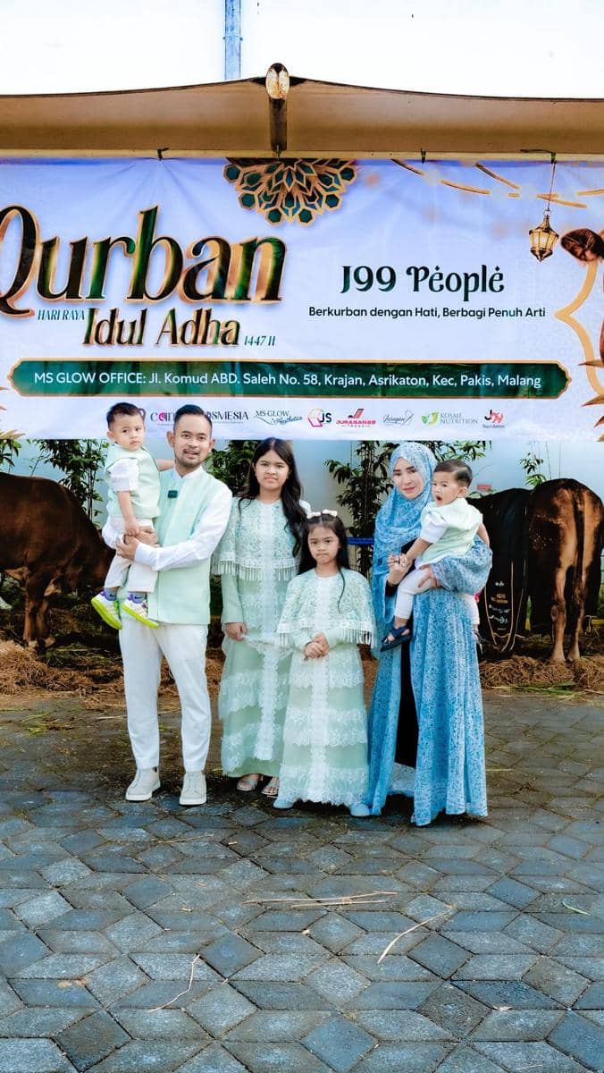 Sebuah keluarga berpose di depan spanduk acara Qurban Idul Adha 1447 H di Malang, mengenakan busana serasi berwarna hijau dan biru.