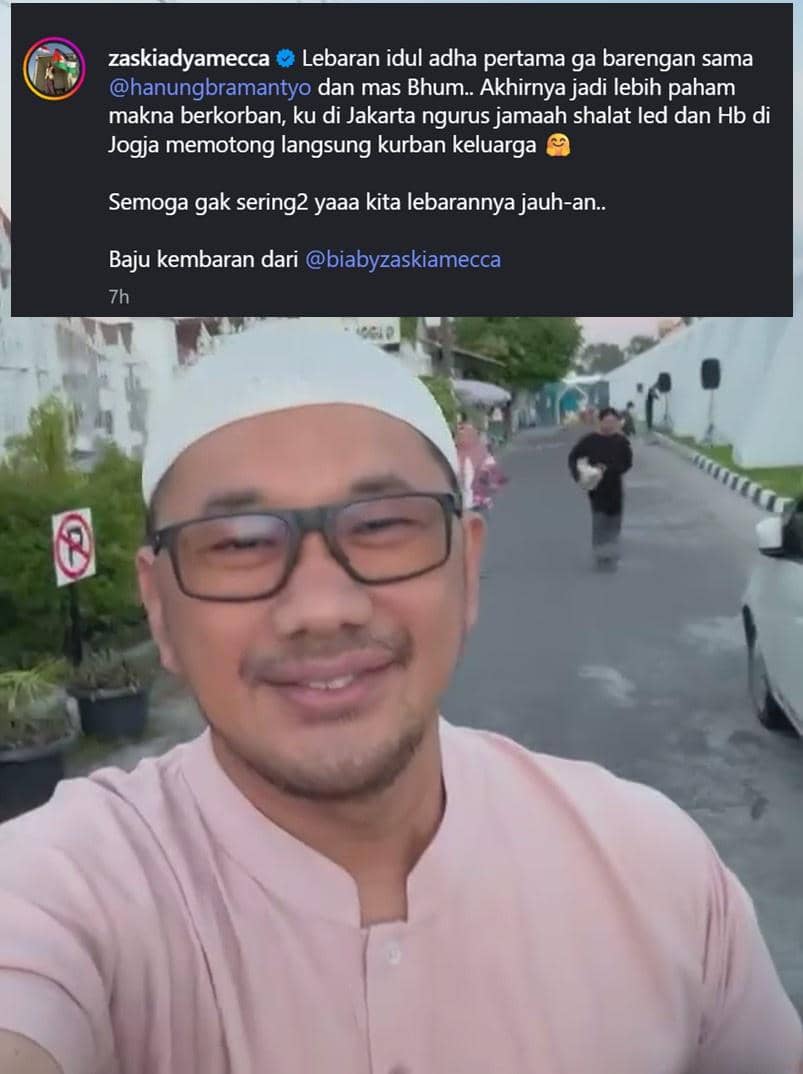 Seorang pria mengenakan peci putih dan kemeja berwarna merah muda tersenyum di jalan dengan latar belakang orang berjalan dan kendaraan terparkir.