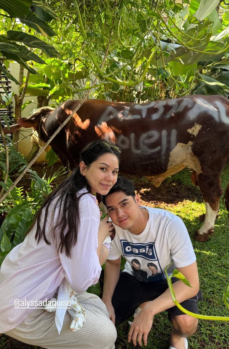 Sepasang orang berpose di taman dengan seekor sapi kurban berwarna cokelat di belakang mereka saat perayaan Idul Adha 2026.