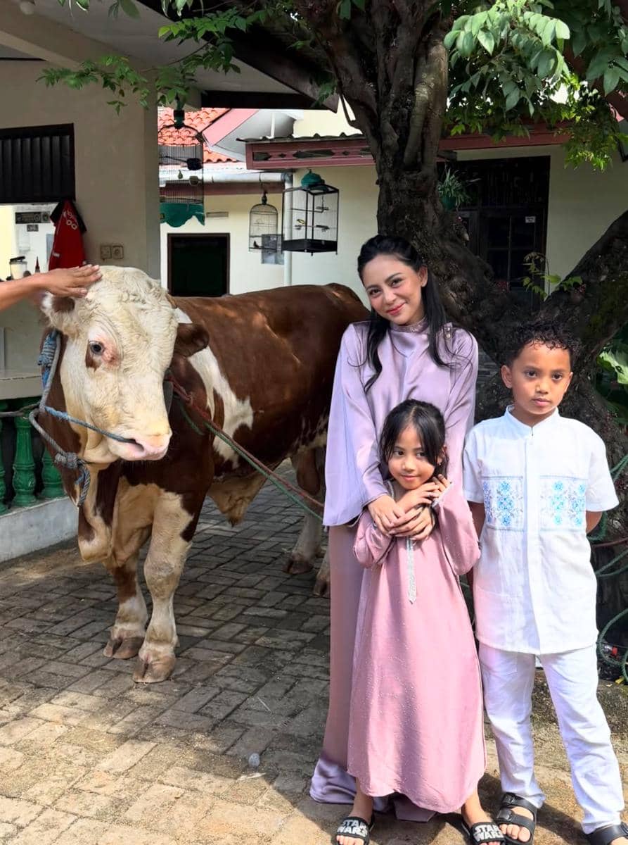 Seorang wanita bersama dua anak kecil berpose di samping sapi kurban berwarna cokelat putih di halaman rumah saat Idul Adha.
