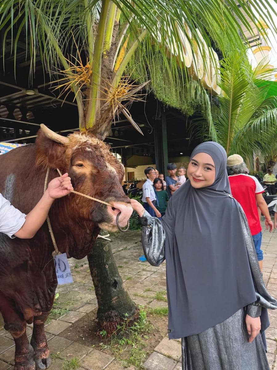 Seorang wanita berhijab abu-abu berpose di samping seekor sapi kurban berwarna cokelat di area terbuka dengan pepohonan kelapa.