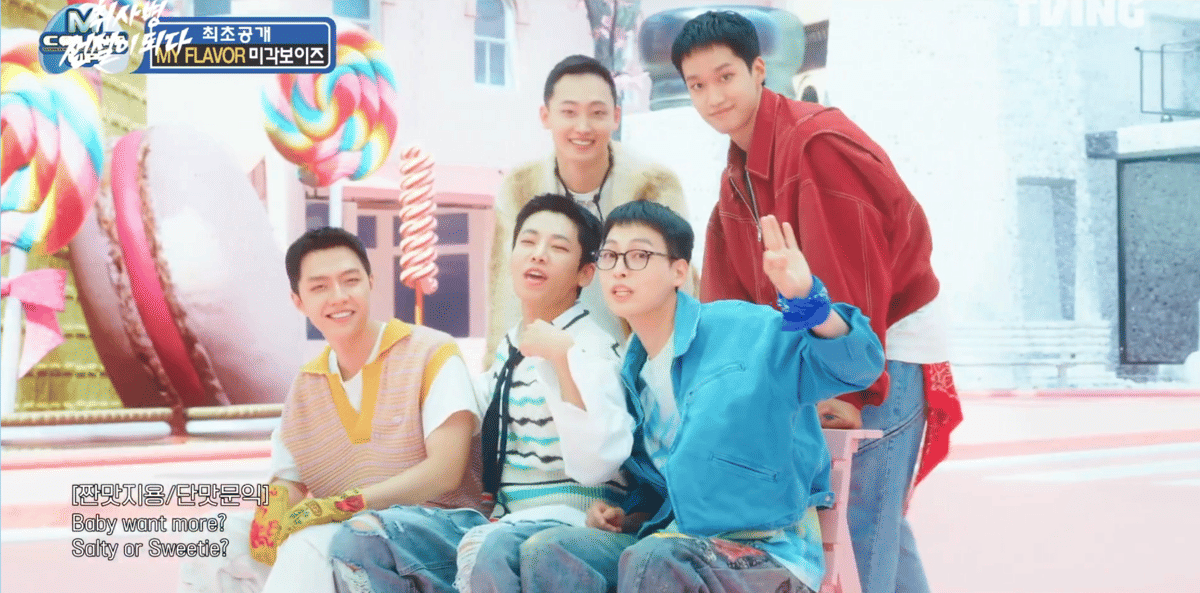 Lima anggota Migak Boys berpose ceria di set berwarna pastel dengan latar permen raksasa dalam cuplikan video musik My Flavor.