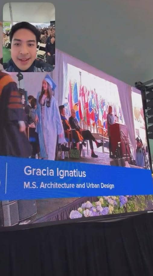 Seorang perempuan mengenakan toga biru berjalan di panggung wisuda dengan layar besar menampilkan namanya, sementara seorang pria terlihat di video call di pojok layar.