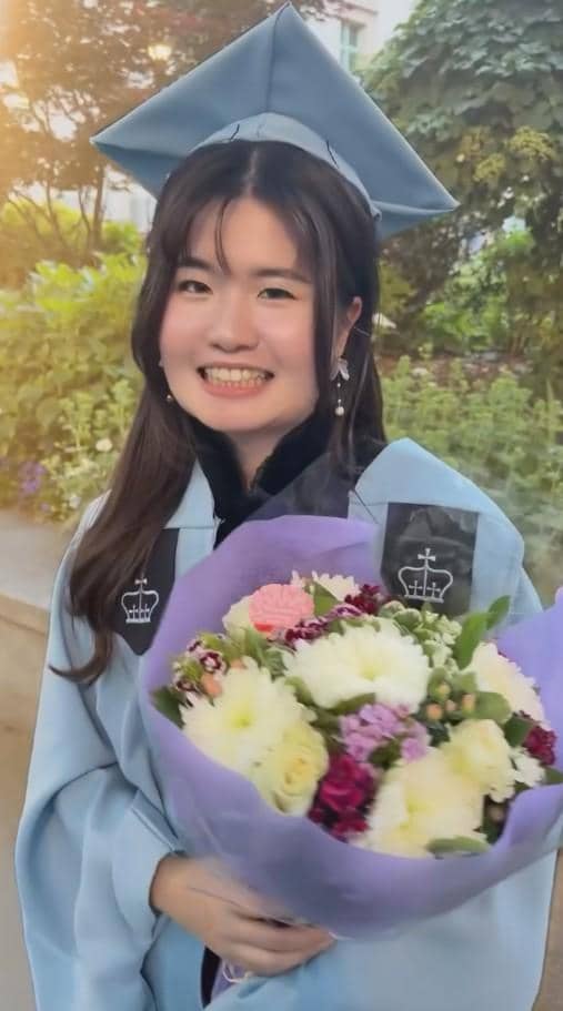 Seorang perempuan mengenakan toga wisuda berwarna biru muda sambil memegang buket bunga berwarna ungu di luar ruangan.