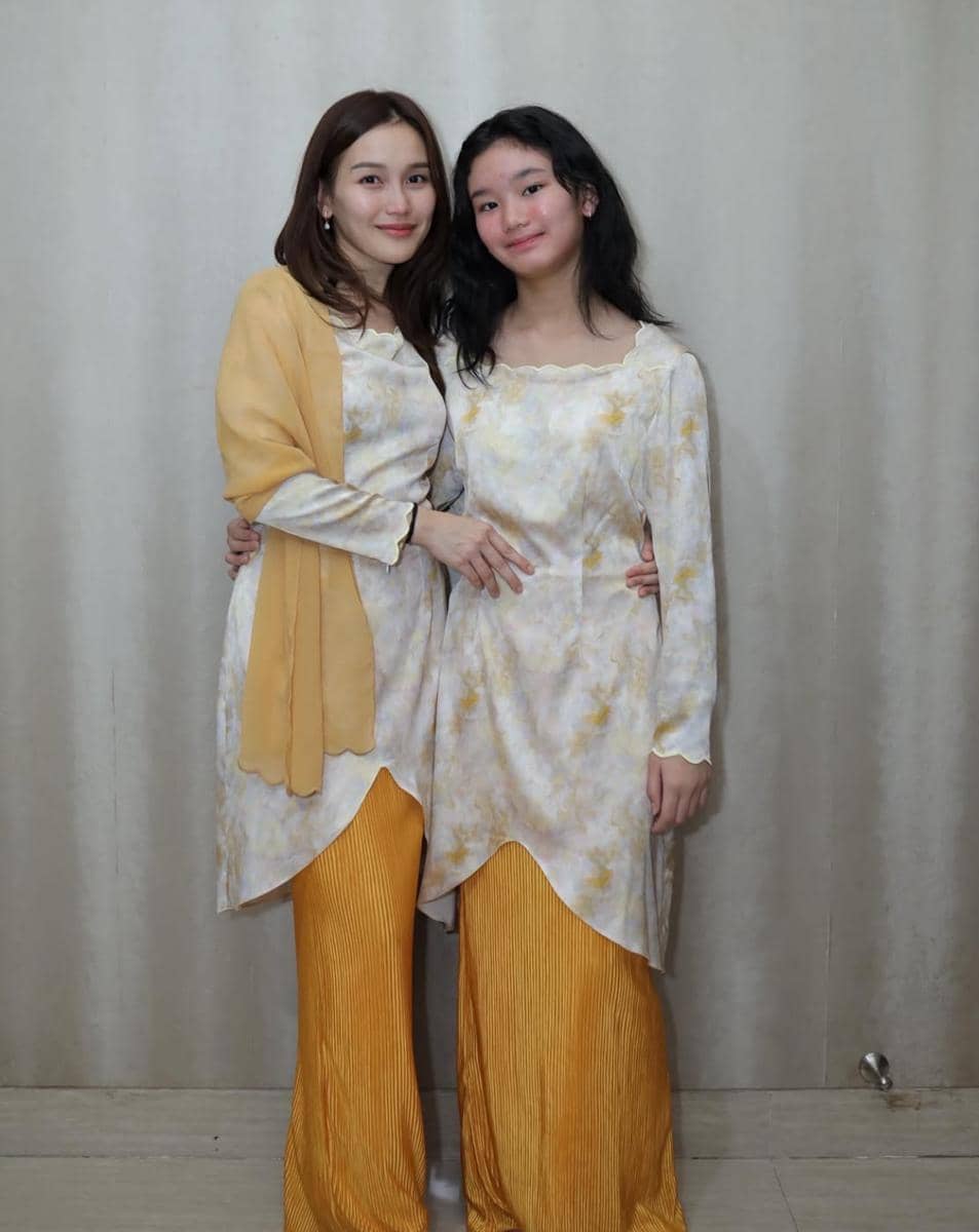 Dua perempuan mengenakan busana senada berwarna kuning dan putih berdiri berdampingan sambil tersenyum di depan latar polos.