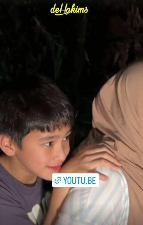 Seorang anak laki-laki tersenyum sambil memegang bahu seseorang berhijab di suasana malam dengan tulisan deHakims di atasnya.