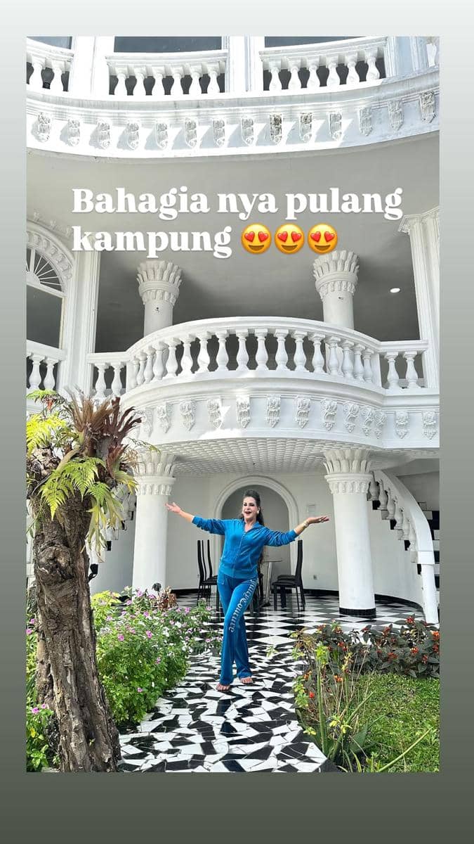 Seorang wanita berpose ceria di depan bangunan bergaya klasik berwarna putih dengan taman hijau dan lantai berpola hitam putih.