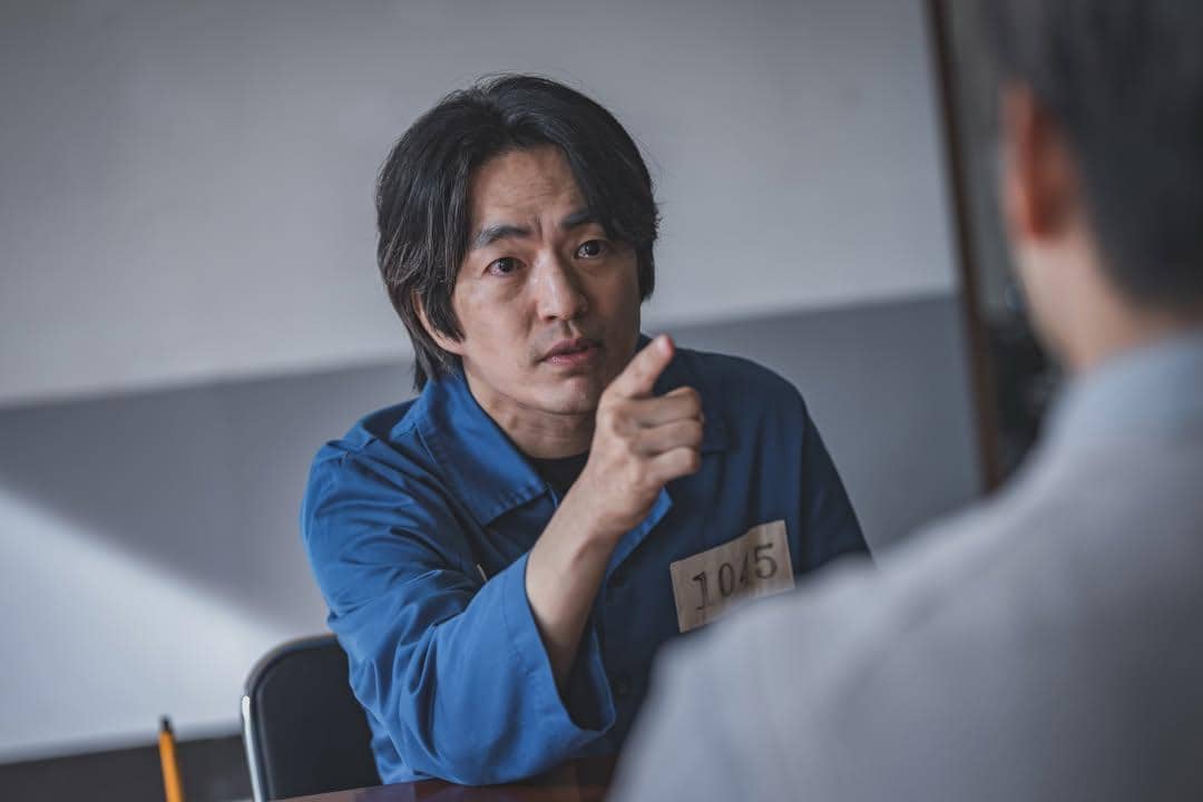 Seorang pria berbaju tahanan biru dengan nomor dada sedang menunjuk seseorang di depannya dalam adegan drama The Scarecrow.