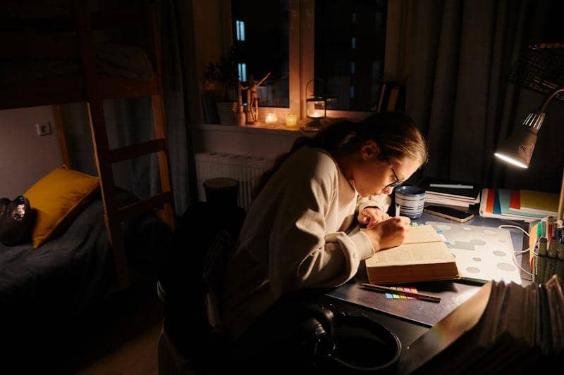 Seorang perempuan belajar di meja dengan pencahayaan lampu malam, dikelilingi buku dan alat tulis di kamar yang hangat.