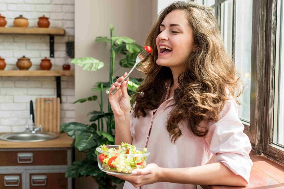 Seorang wanita tersenyum sambil memegang mangkuk salad dan akan memakan tomat ceri dengan garpu di dapur yang terang.