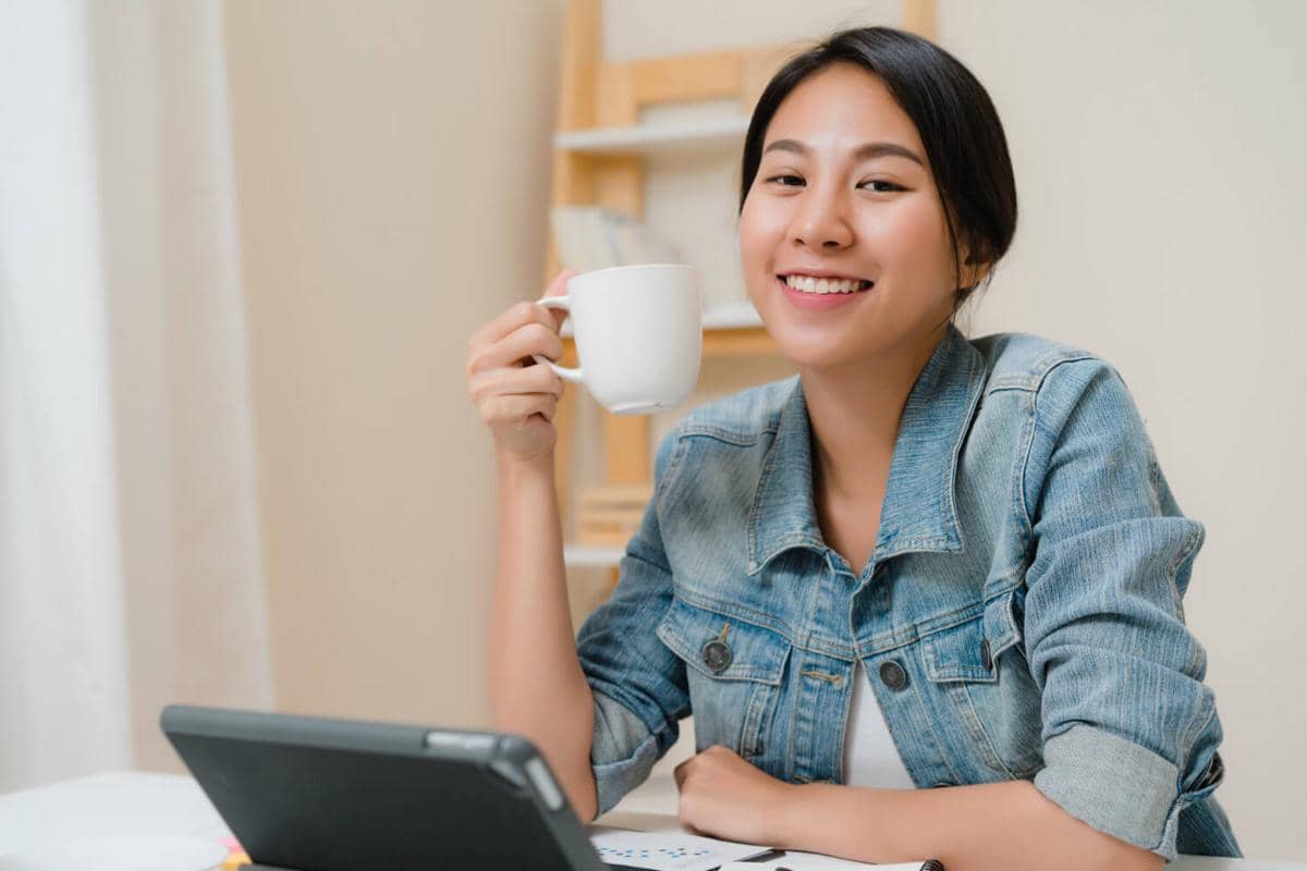 Seorang perempuan tersenyum sambil memegang cangkir kopi di tangan, duduk di depan tablet di meja kerja dengan suasana santai.