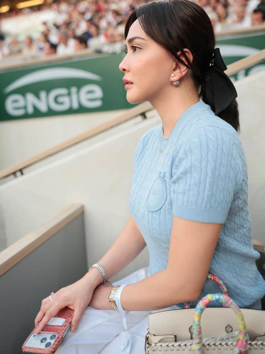 Seorang perempuan mengenakan atasan rajut biru muda duduk di tribun menonton pertandingan tenis di Paris dengan suasana ramai di sekitarnya.