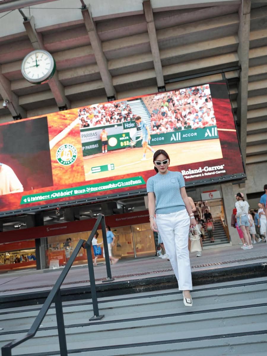 Seorang wanita mengenakan kacamata hitam, atasan biru, dan celana putih berjalan di area stadion tenis Roland-Garros di Paris.
