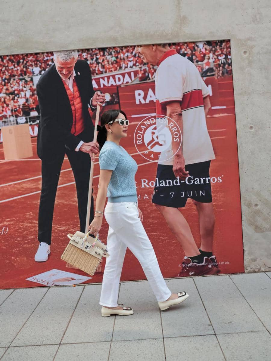 Seorang wanita mengenakan atasan biru dan celana putih berjalan di depan poster turnamen tenis Roland-Garros di Paris.