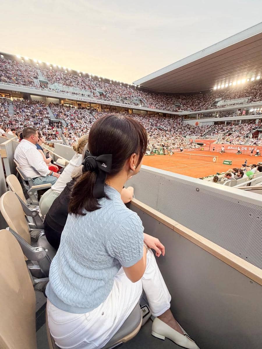 Seorang wanita berbusana biru muda dan putih duduk di tribun stadion tenis yang penuh penonton saat pertandingan di Paris.