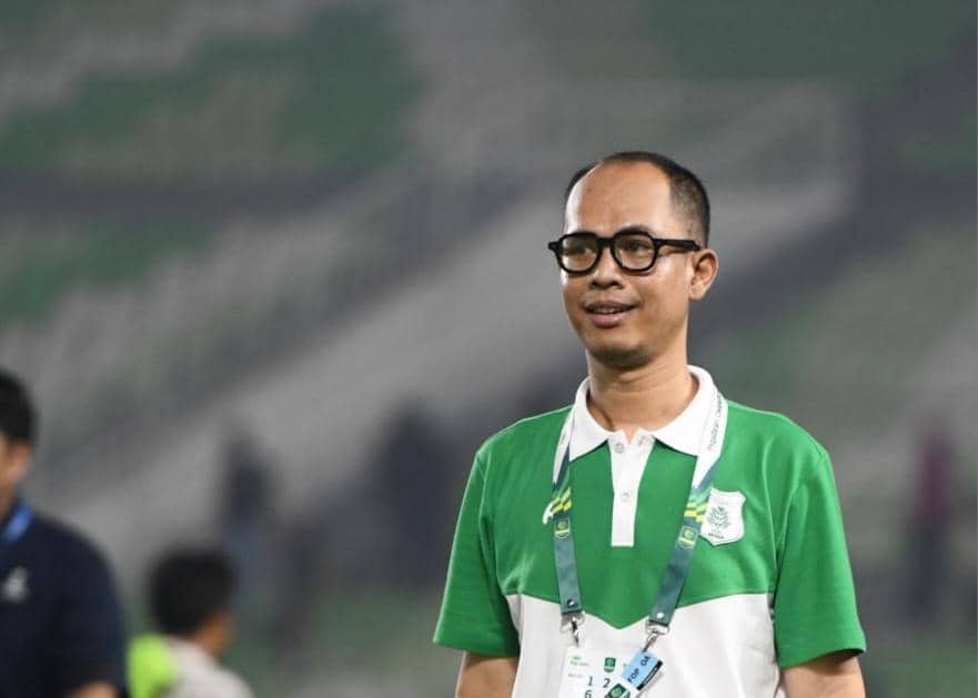 Seorang pria berkacamata mengenakan kaus hijau putih dengan logo PSMS berdiri di area stadion dengan latar tribun penonton.