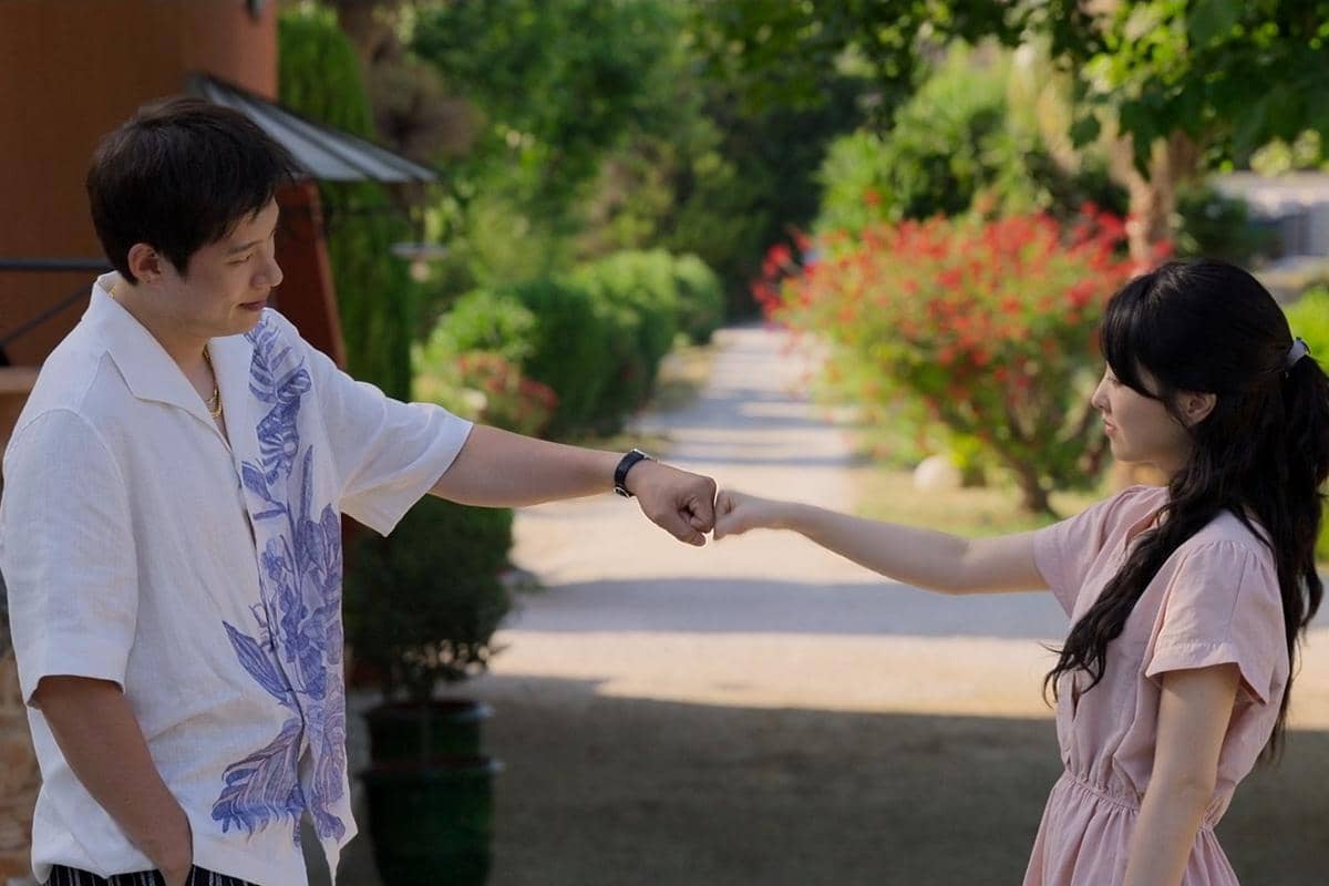 Dua karakter dalam drama Korea Gold Land saling menyentuh kepalan tangan di jalan taman yang dikelilingi pepohonan hijau.