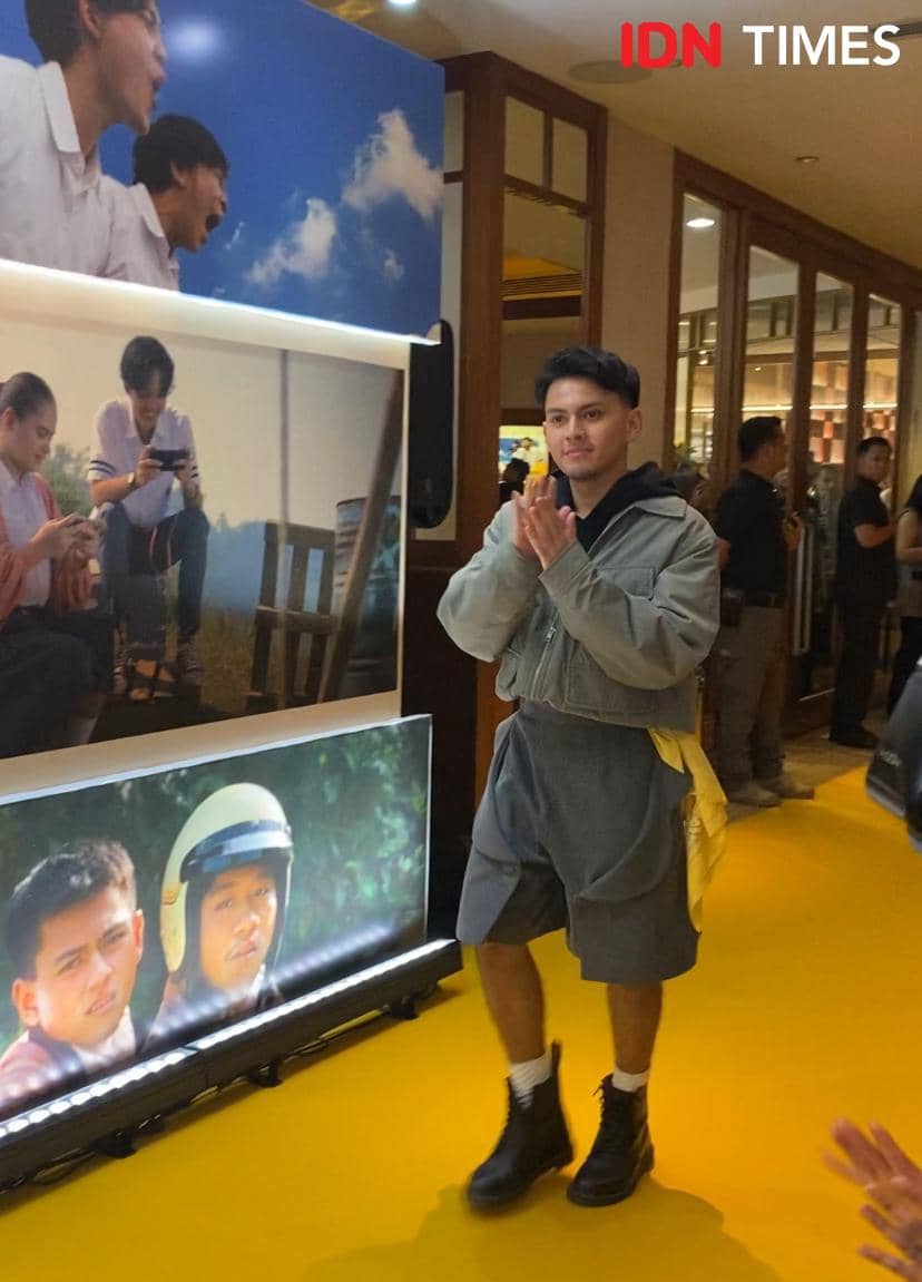 Seorang pria berjalan di karpet kuning acara pemutaran film Nobody Loves Kay, mengenakan jaket abu-abu dan celana pendek dengan sepatu boot hitam.