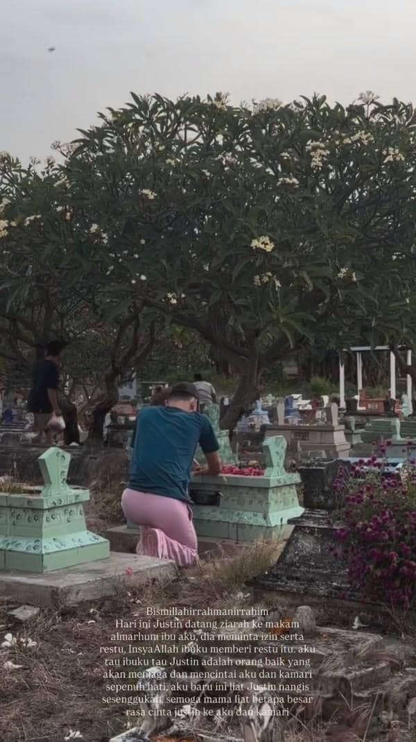 Seorang pria dan wanita berziarah di pemakaman, duduk di depan nisan dengan taburan bunga di bawah pohon kamboja yang rimbun.