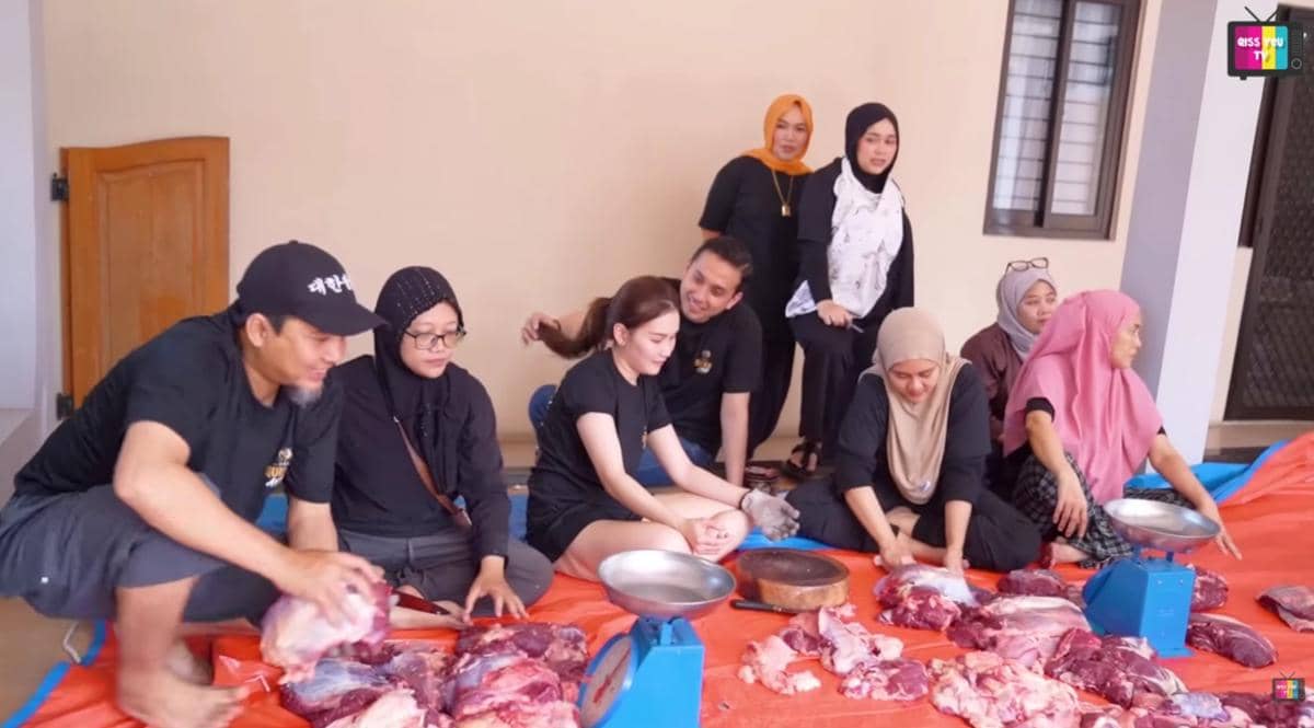 Sekelompok orang mengenakan pakaian santai sedang memotong dan menimbang daging sapi kurban di atas terpal oranye di halaman rumah.