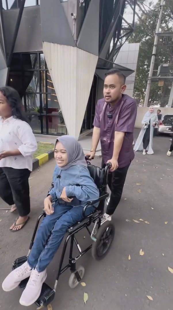 Seorang pria mendorong kursi roda berisi perempuan berhijab yang tersenyum di area luar gedung modern bersama beberapa orang lain.