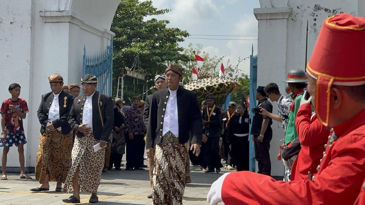 Para abdi dalem mengenakan busana adat Jawa berjalan dalam prosesi Grebeg Besar Idul Adha di bawah gerbang keraton dengan hiasan gunungan dan bendera merah putih.