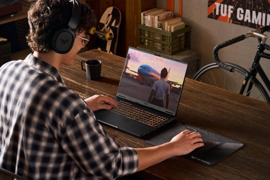 Seseorang mengenakan headphone bermain game di laptop ASUS TUF Gaming dengan pencahayaan keyboard oranye di atas meja kayu.