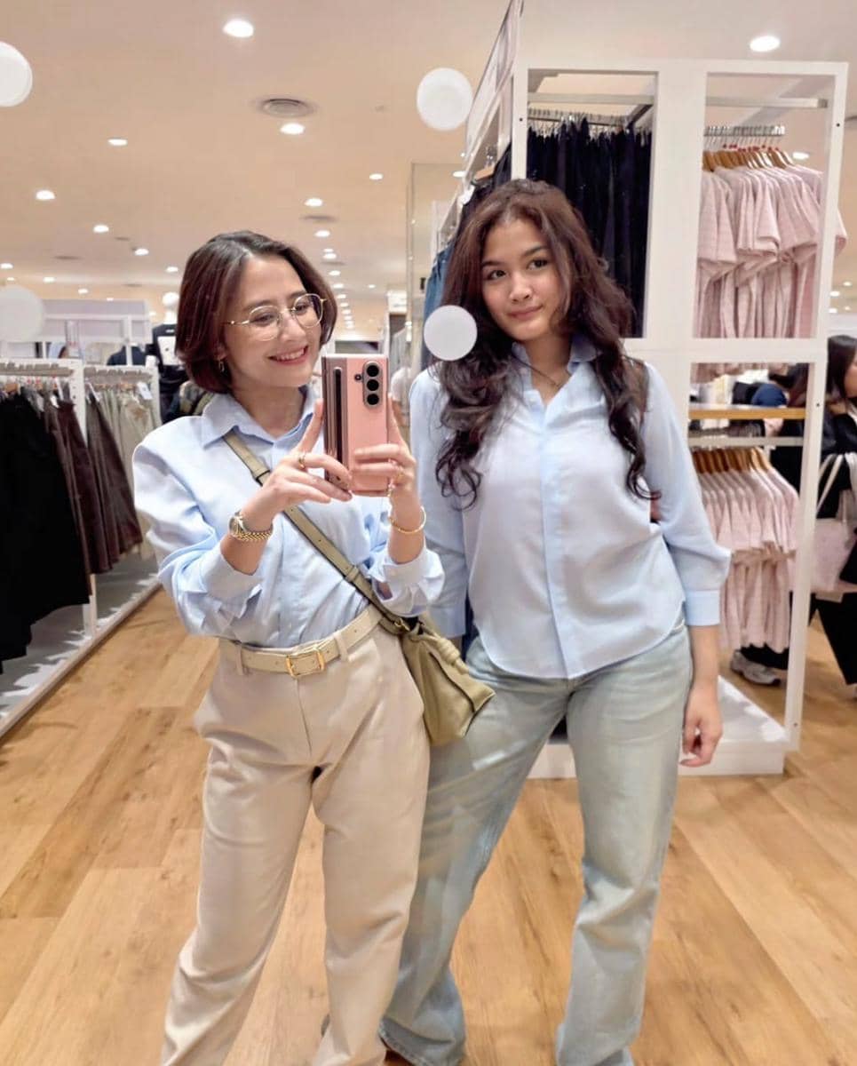 Dua perempuan berpose di depan cermin di dalam toko pakaian, mengenakan kemeja biru muda dan celana berwarna terang.