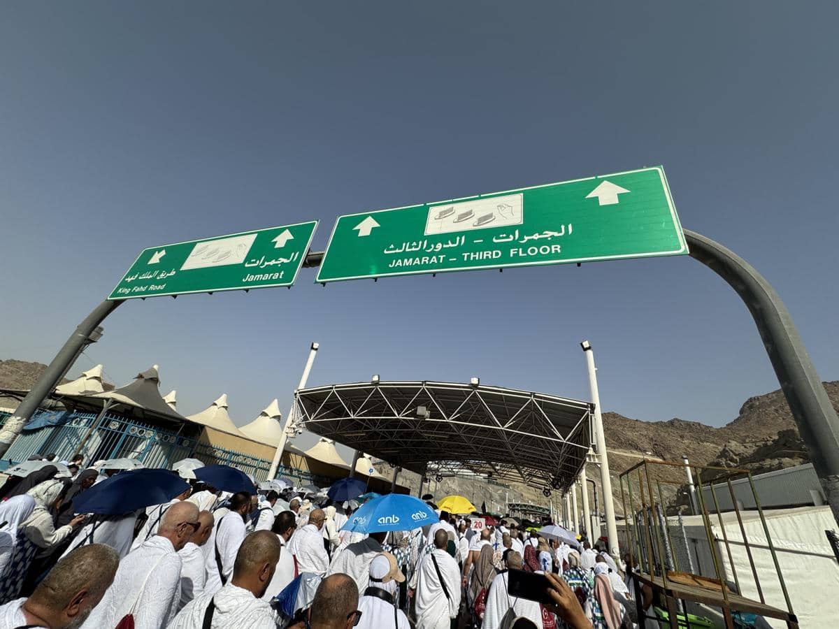 Rombongan jamaah haji berjalan menuju area Jamarat di Mina, Makkah, di bawah papan petunjuk arah berwarna hijau pada siang hari yang cerah.