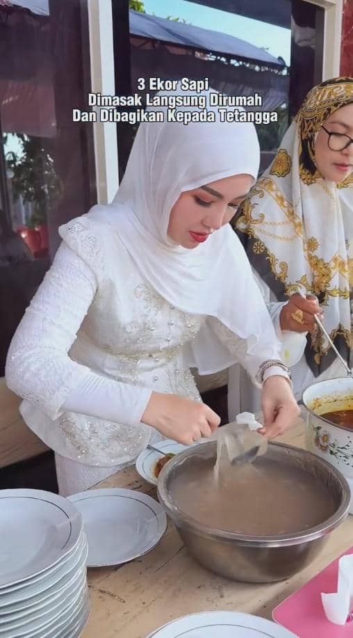 Seorang wanita berhijab putih sedang menyiapkan makanan dalam wadah besar di rumah, bersama wanita lain mengenakan hijab bermotif.