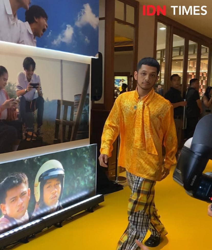 Seorang pria berjalan di karpet kuning acara pemutaran film Nobody Loves Kay dengan mengenakan kemeja oranye dan celana motif kotak.