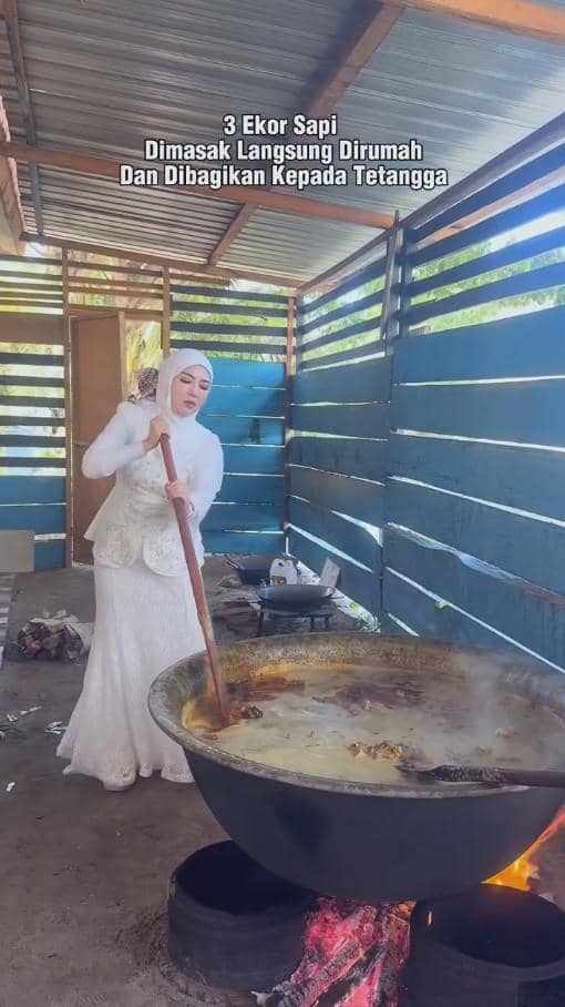 Seorang wanita berhijab mengenakan pakaian putih sedang mengaduk masakan besar di kuali di dapur terbuka dengan api unggun di bawahnya.