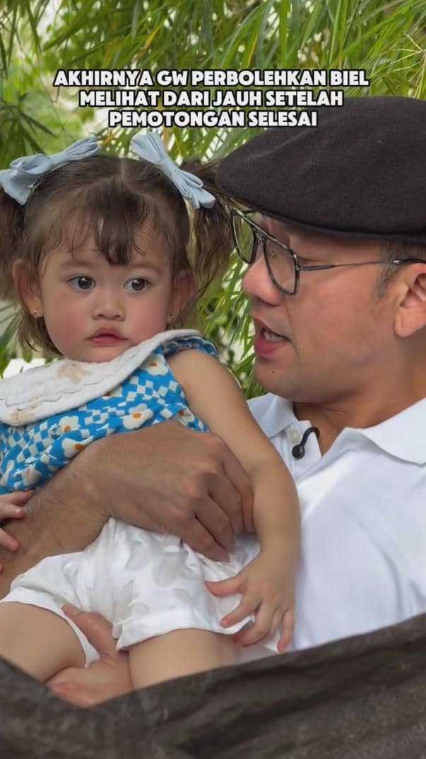 Seorang pria mengenakan topi dan kacamata memeluk anak kecil bergaun biru bermotif bunga dengan pita di rambutnya di luar ruangan.