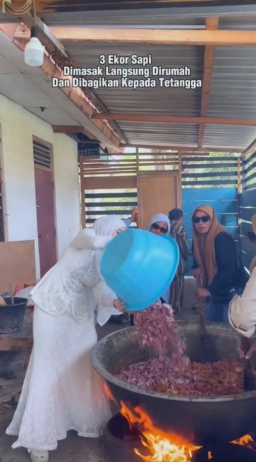 Seorang wanita berpakaian putih menuangkan daging sapi ke dalam wajan besar di atas api, dibantu beberapa orang di dapur terbuka.